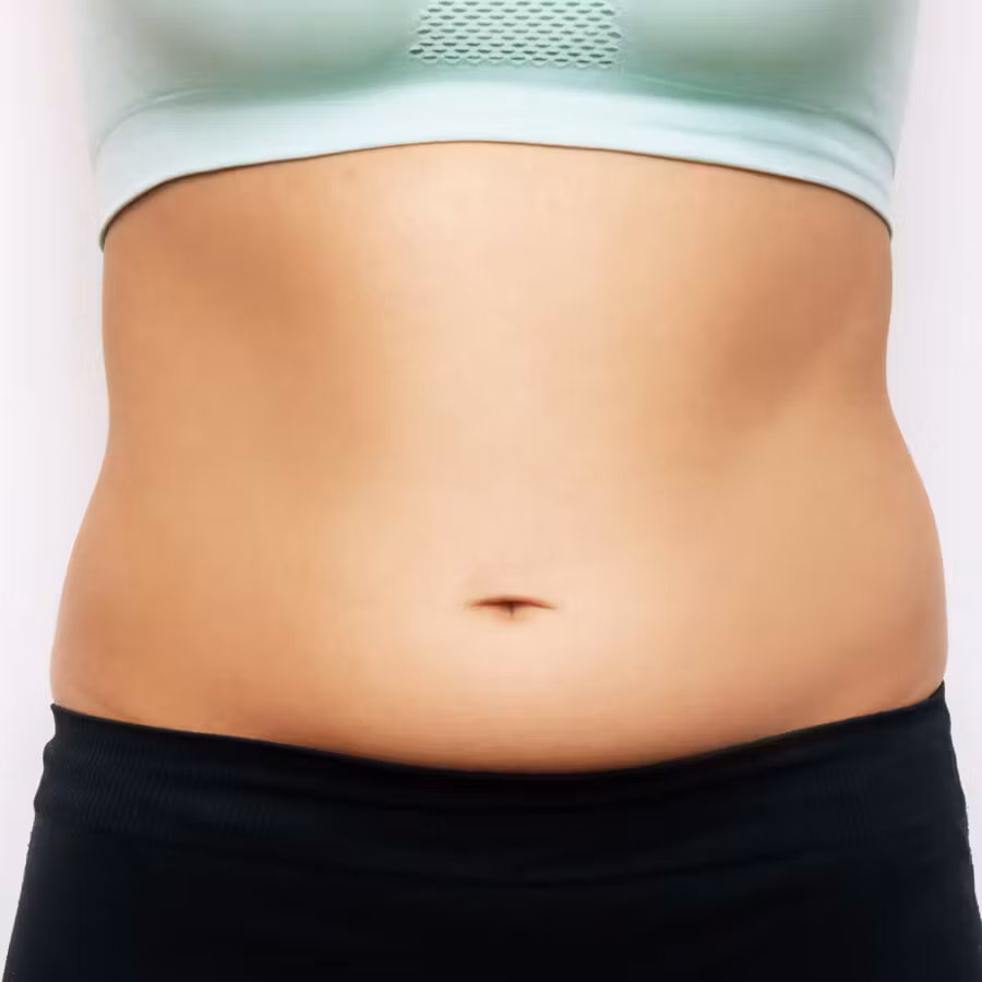A close-up view of a person’s abdomen, showing the midsection between a light-colored sports bra and black athletic bottoms against a plain background.